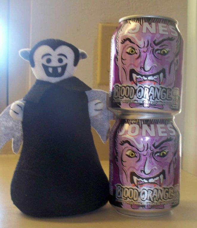 Photo of a Little Vampire plush toy posing next to cans of Jones Blood Orange Soda