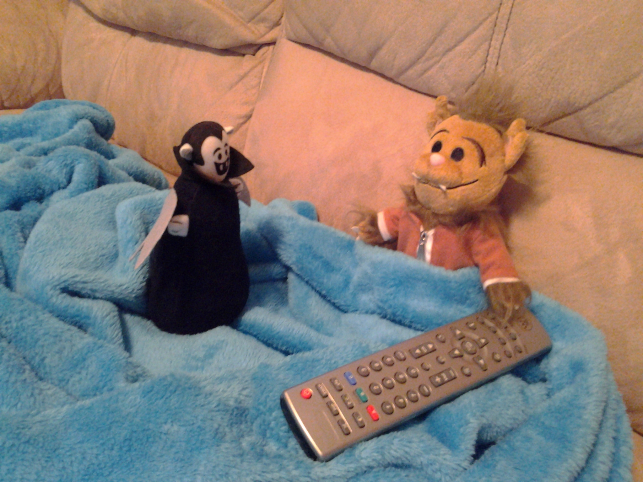 Photo by Linda Ratliff of a Wolfie plush toy and a Little Vampire plush toy sitting on a couch with a blanket and a television remote control