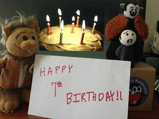 Photo of a Wolfie plush toy, a Little Vampire plush toy, and a Little Vampire wooden doll posed around a sign reading “Happy 7th Birthday!!” all standing in front of a birthday cake wih seven lit candles. The Little Vampire plush toy and wooden doll are standing on a box with a Little Vampires sticker on it.
