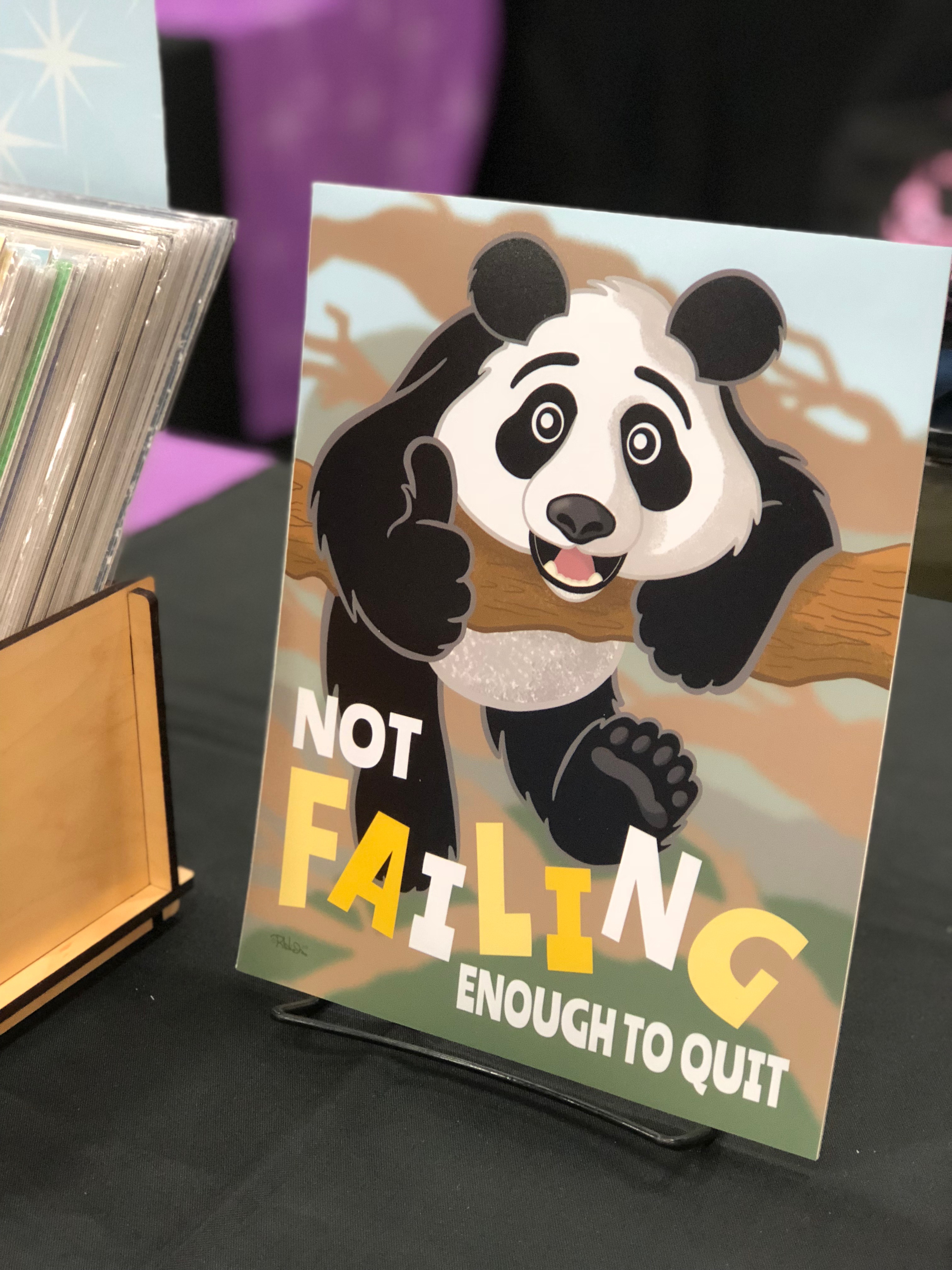 Photo of an art print depicting a panda barely hanging onto a branch, but giving a thumbs-up, captioned “Not Failing Enough to Quit!