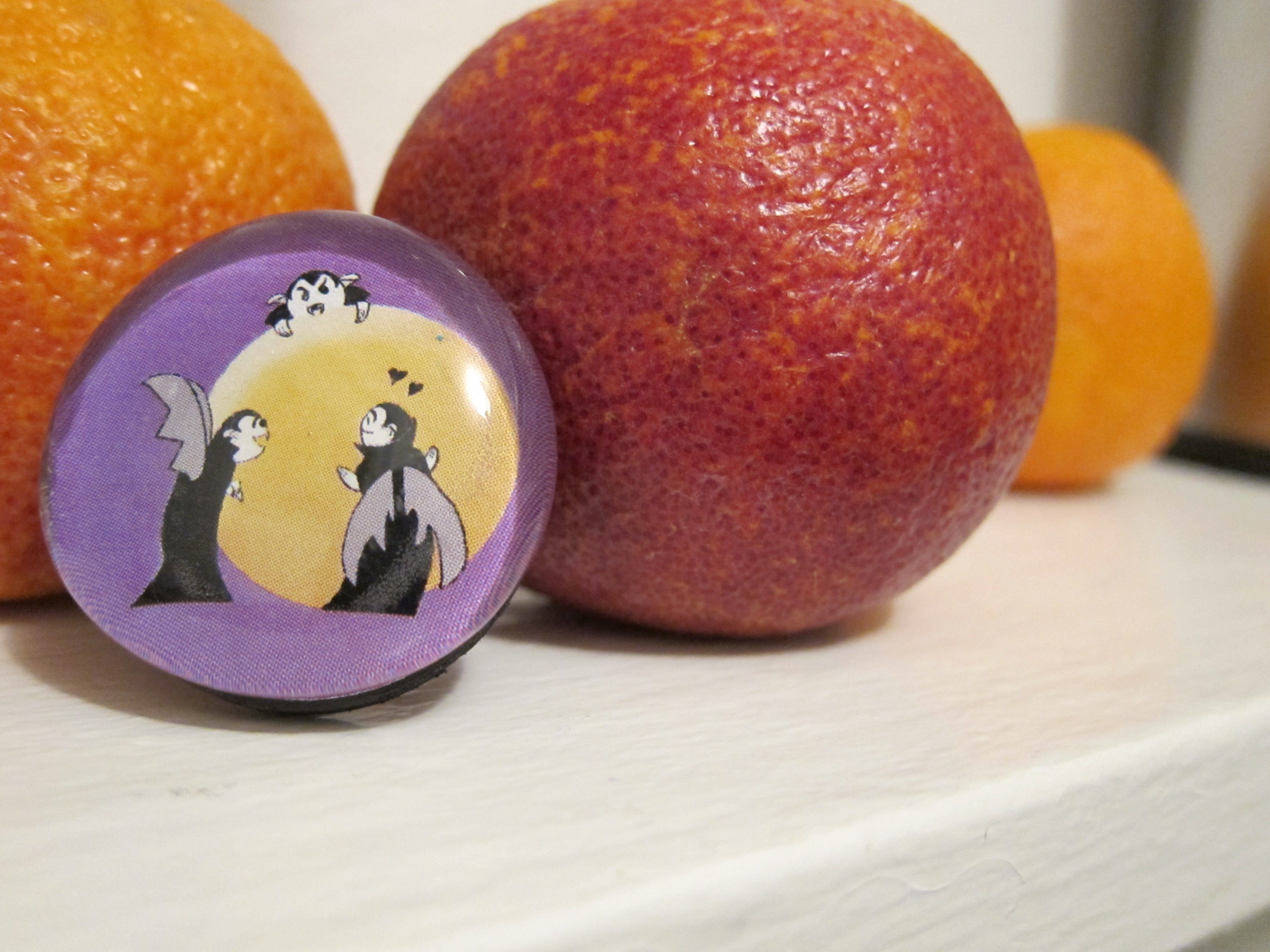 Photo of a Little Vampires glass refrigerator magnet depicting Little Vampirea and a blood orange. The magnet is arranged with a few real blood oranges.