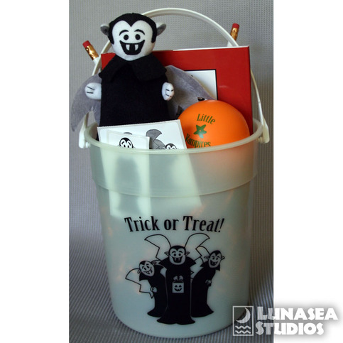 Photo of a glow-in-the-dark Little Vampires trick-or-treat pail. The pail contains two Little Vampires wooden pencils, a blood orange stress ball, a Little Vampires hardcover book, and Little Vampires temporary tattoos.