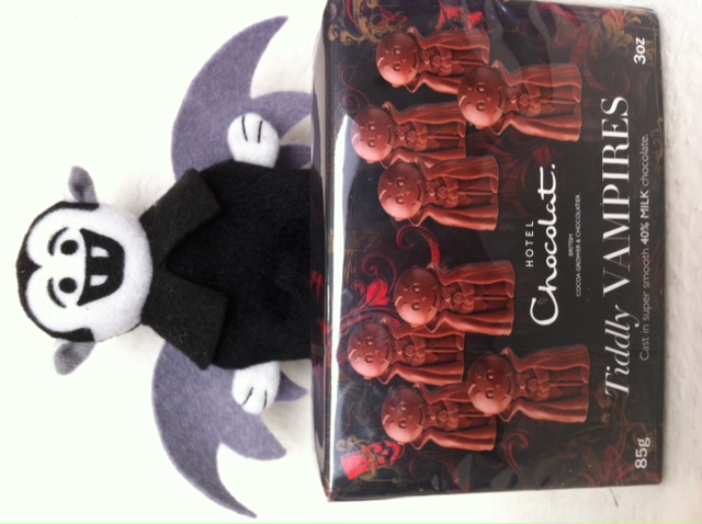 A photo of a Little Vampire plush toy posing behind a box of chocolate Tiddly Vampires candy
