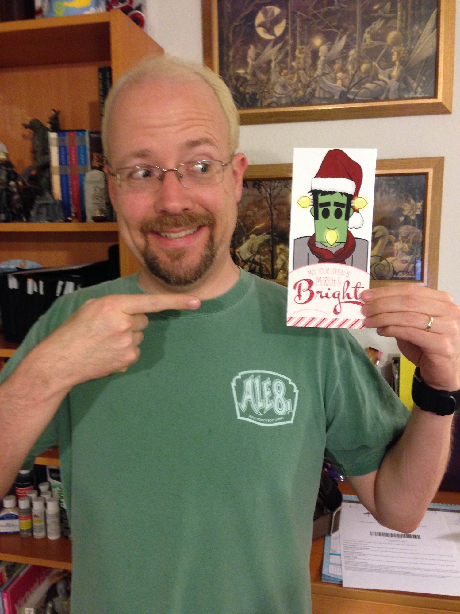 Photo of James Hicks holding a holiday card and pointing to it. The card depicts Frank wearing a Santa hat, with lightbulbs on the sides of his head and in his mouth. The card reads “May Your Holiday Be Merry and Bright”